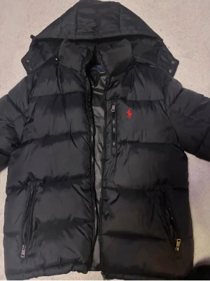 Svart pufferjacka från Polo Ralph Lauren - Svart pufferjacka från Polo Ralph Lauren med röd broderad logga på bröstet. Jackan har huva, två stora fickor med dragkedja och kraftig dragkedja framtill. Materialet är glansig och jackan är vadderad för extra värme. Perfekt för kalla dagar.
