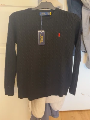 Svart kabelstickad tröja Polo Ralph Lauren - Kabelstickad svart tröja från Polo Ralph Lauren med klassisk röd logga broderad på bröstet. Tröjan har rund halsringning och långa ärmar, perfekt för dig som gillar stilrena och tidlösa plagg. Passar till allt och är superenkel att matcha.