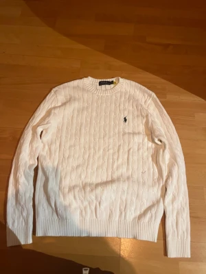 Vit kabelstickad tröja Polo Ralph Lauren - Vit kabelstickad tröja från Polo Ralph Lauren med klassisk rund hals och broderad marinblå logga på bröstet. Tröjan har ribbade muddar vid ärmslut och nederkant, och är tillverkad i mjuk bomull. Perfekt för dig som gillar stilrena och tidlösa plagg. Den är i storlek s men är stor i sin storlek