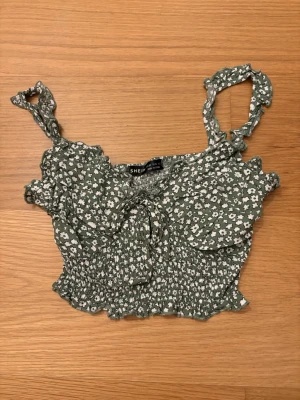 Grön blommig croppad topp från SHEIN, storlek XS - Supersöt grön croppad topp från SHEIN i storlek XS med vita små blommor, smockad midja och volangkant. Axelbanden är knutna och toppen har en liten rosett framtill. Perfekt för varma dagar och ger en riktigt somrig vibe.