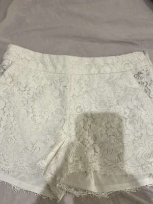 Vita spets-shorts från Amisu - Säljer ett par vita shorts från Amisu i storlek 36. De är täckta med blommig spets och har en diskret dragkedja i sidan. Shortsen har  fransiga kanter nertill för en extra snygg detalj. Perfekta till sommaren! 