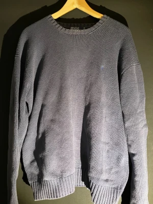 Mörkblå stickad tröja Polo Ralph Lauren - Klassisk mörkblå stickad tröja från Polo Ralph Lauren i 100% bomull. Tröjan har rund halsringning, ribbade muddar och diskret broderad logga på bröstet. Skriv vid frågor