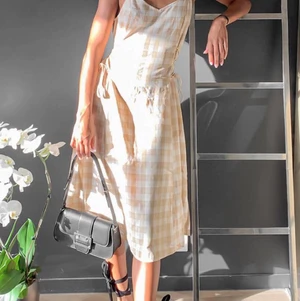 Vacker klänning ifrån Vila - Plagget är vadlångt på mig som är 170 och är i en ljus beige färg och har ett jättefint rutigt mönster som är så fint i sommar. Knytning i sidan som man justerar själv vilket ger en fin figur.  Köpt i mars men aldrig använt pågrund av för stor passform. Köparen står för frakt vilket ligger på 66kr.