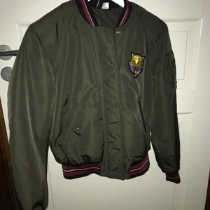 H&M bomberjacka strl 32 - Grön bomber jacka med röda detaljer och en liten tiger på. Jackan kommer från H&M och är i bra skick. Storlek 32