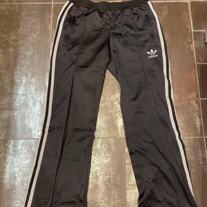 Trendiga Adidas track pants - Ett par supersnygga och trendiga track pants från adidas. Byxorna är svarta med glittersilvriga streck och en logga på vänster ben. Fickor med dragkedja på båda sidor och dragkedja längst ner på byxorna.