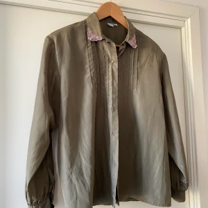 Blus - Blus i militärgrönt färg och broderade, rosa/lila detaljer. 🌸 Mindre axelvaddar som enkelt går att ta bort. OBS! Saknas en knapp längst ner, dock inget som syns när blusen är knäppt. **ev frakt ingår i priset!