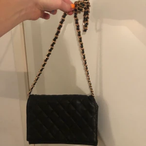 Svart liten väska  - Detta är en väska i svart fake skinn. Bandet är i guld och svart. Den är En knapp är lite dålig men den funka