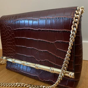 Mörkröd väska med guldig kedja  - En mörkröd väska med guldiga detaljer och kedja. Materialet är fake skinn. Fint skick.