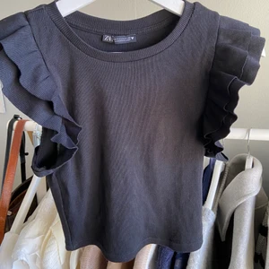 Volang tröja Zara - Svart ribbad t-shirt från Zara med volang ”ärmar”. Köpte den i slutet på sommaren men har använt den 2 gånger så säljer vidare den, den är som i nytt skick!☺️