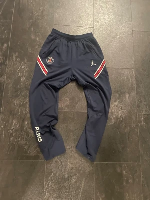 Blå PSG x Jordan träningsbyxor - Mörkblå träningsbyxor från Jordan i samarbete med PSG. Byxorna har vita och röda detaljer vid fickorna, PSG-logga på höger sida och Jumpman-logga på vänster. Texten 'PARIS' finns tryckt på vänster ben. Perfekta för fotboll eller gym. Har också två små hål vid båda fickorna.