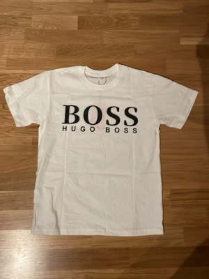 Vit Hugo Boss t-shirt med logga - Snygg vit t-shirt med stort svart BOSS tryck på bröstet . Klassisk rund hals och korta ärmar. Tillverkad i mjuk bomull för en skön känsla och enkel stil som funkar till allt.