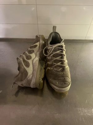 Nike Air Vapormax Plus vita sneakers - Nike Air Vapormax Plus sneakers i vitt och grått med genomskinlig sula och ikoniska vågiga detaljer på sidan. Ovandelen är i textil och syntet, snörning framtill och dragflik bak. Snygg chunky design som sticker ut.