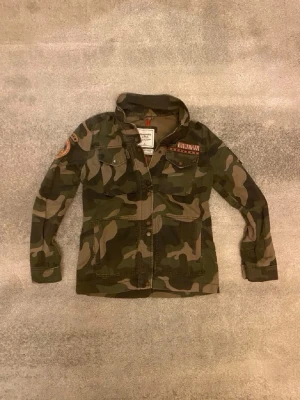 Camo vindjacka från Abercrombie & Fitch - Snygg vindjacka i camomönster från Abercrombie & Fitch. Jackan har två bröstfickor med knappar, patchdetalj på ärmen och dekorativa märken vid bröstet. Tillverkad i slitstark bomullsmix och har en avslappnad passform med krage.