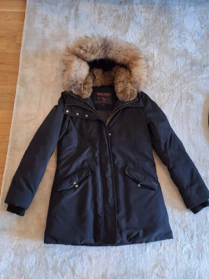 Svart parkas från Woolrich med päls - Snygg svart parkas från Woolrich med stor fluffig pälskant på huvan och quiltat foder. Jackan har dragkedja, knappar och två snedställda fickor framtill. Perfekt för kalla dagar och riktigt varm med klassisk look.