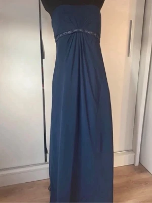 Marinblå axelbandslös långklänning - Säljer en marinblå axelbandslös långklänning med dekorativt band under bysten och lätt rynkning framtill. Klänningen har en elegant och enkel siluett. Dragkedja i ryggen för enkel påklädning.