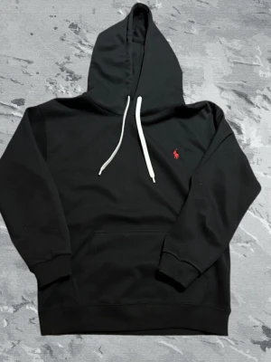 Svart hoodie från Polo Ralph Lauren - Svart hoodie från Polo Ralph Lauren med klassisk huva och dragsnören. På bröstet finns den ikoniska röda Polo-loggan broderad. Hoodien har ribbade muddar och är tillverkad i mjukt material som känns skönt mot huden. Perfekt för en avslappnad och stilren look.