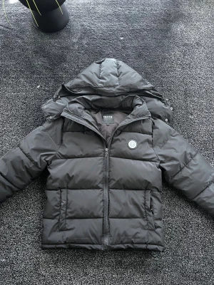 Trapstar vinterjacka - Snygg svart pufferjacka från Trapstar med avtagbar huva och dragkedja framtill. Jackan har Trapstar-logga på bröstet och stor broderad text på ryggen. Perfekt för kalla dagar och riktigt streetwear-vibe. Storlek L, skick 9/10, pris går att diskutera✅