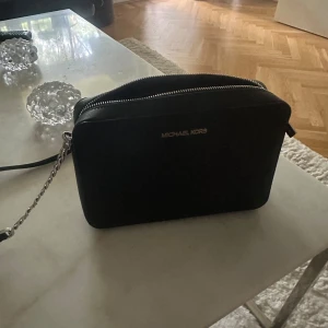 Svart axelväska från Michael Kors - Snygg svart axelväska från Michael Kors i stilren, rektangulär modell. Väskan har silverfärgade detaljer, kedja på axelremmen och dragkedja upptill. Tillverkad i skinn och har två fack inuti för smidig organisering.