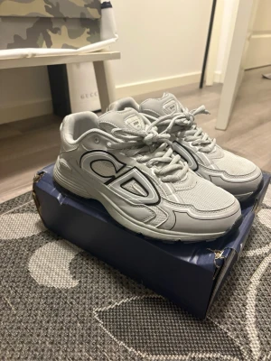 Gråa Dior B30 sneakers - Säljer ett par gråa Dior B30 sneakers med stora CD-loggor på sidorna. Skorna har en sportig och chunky design med mesh och syntetmaterial, samt snörning framtill. Perfekta för dig som gillar exklusiva och trendiga sneakers.