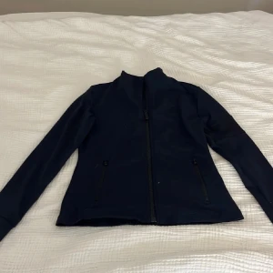 Maya delorez ridjacka, mörkblå - Mörkblå tunn ridjacka med hel dragkedja framtill och två fickor med dragkedja. Jackan är långärmad och har en hög krage. Det är en diskret logga upptill nacken.Materialet känns stretchigt. Perfekt till en vår eller sommar dag i stallet eller ett ridpass.