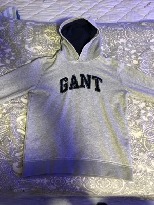 Grå hoodie från GANT med tryck - Snygg grå hoodie från GANT med svart broderad logga på bröstet. Mjuk insida och fodrad huva i mörkblått. Perfekt för dig som gillar klassisk och sportig stil. Långa ärmar och ribbade muddar.