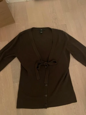 Brun kofta med knytband från H&M - Mysig brun kofta från H&M med knytband i midjan och knappar framtill. Djup V-ringning och långa ärmar. Perfekt att slänga på sig över en topp för en chill och stilren look!!🤎🤎