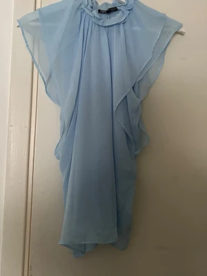 Ljusblå blus med volang från Zara - Säljer en ljusblå blus från Zara med volangdetaljer och hög krage. Blusen är ärmlös och har ett luftigt, flowigt material som känns riktigt skönt. Perfekt för dig som gillar en feminin och trendig look. Hör av er för prisförslag elelr fler bilder❤️