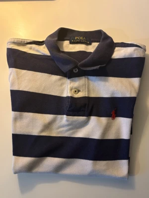 Randig piké från Polo Ralph Lauren - Klassisk pikétröja från Polo Ralph Lauren med breda marinblå och vita ränder. Tröjan har krage, knappar framtill och den ikoniska röda logotypen broderad på bröstet. Tillverkad i bomull för en bekväm och avslappnad stil.