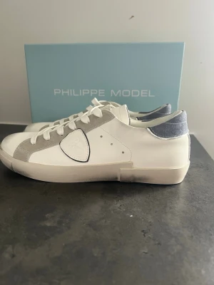 Vita Philippe Model sneakers i skinn - Stilrena vita sneakers från Philippe Model med grå mockadetaljer och blågrå häl. Klassisk låg modell med vita skosnören och logotyp på sidan. Ovandel i skinn och mocka, sula i gummi. Perfekta för en clean och modern look.