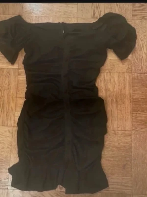 Svart offshoulder kortklänning - Trendig svart kortklänning med offshoulder-design och rynkade detaljer längs hela klänningen. Klänningen har en kroppsnära passform och korta puffärmar som ger en snygg siluett. Perfekt för dig som vill ha en stilren och modern look.