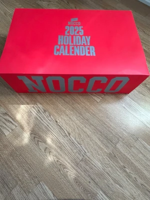 NOCCO 2025 Holiday Calender - Stor röd box från NOCCO med texten '2025 Holiday Calender' i vitt.3D-tryck på sidan med NOCCO-loggan. Perfekt för dig som gillar energidrycker och vill ha en unik kalender till högtider.