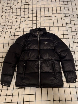 Svart pufferjacka från Prada - Snygg svart pufferjacka från Prada med logga på bröstet och insidan. Jackan har hög krage, två dragkedjefickor och stängs med dragkedja. Perfekt för kalla dagar och ger en stilren look med sin blanka finish och klassiska design.