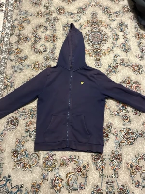 Mörkblå hoodie från Lyle & Scott - Mörkblå hoodie med dragkedja från Lyle & Scott. Klassisk design med huva, kängurufickor och gul broderad logga på bröstet. Perfekt för en avslappnad och stilren look.