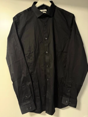 Svart skjorta från Jack & Jones - Klassisk svart skjorta från Jack & Jones Premium med stretchmaterial. Skjortan har lång ärm, knappar framtill och en stilren krage. Perfekt för dig som gillar en enkel och clean look.