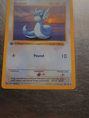 Pokémonkort Dratini 1st Edition  - Säljer ett original Pokémonkort från Base Set med Dratini. Kortet har gul ram, motiv av Dratini mot solnedgång, och klassisk Pokémon-logga på baksidan. Kortet är från 1999 och har samlarnummer 26/102. Perfekt för samlare eller nostalgiker.