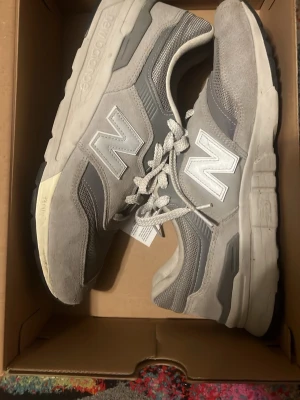 Grå New Balance 997 sneakers - Snygga New Balance 990 sneakers i olika nyanser av grått med klassisk N-logga på sidorna. Ovandelen är i en mix av mocka och mesh för både stil och komfort. Tjock sula för extra support och retrovibbar. Perfekta för dig som gillar streetwear och sportig stil.
