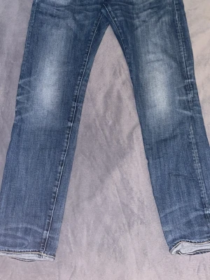 Blå raka jeans med slitningar - Säljer ett par klassiska blå jeans med raka ben och snygga slitningar på framsidan. Jeansen har fem fickor och kontrastsömmar. Materialet är denim i bomull och de har en tidlös tvättad look som passar till det mesta.