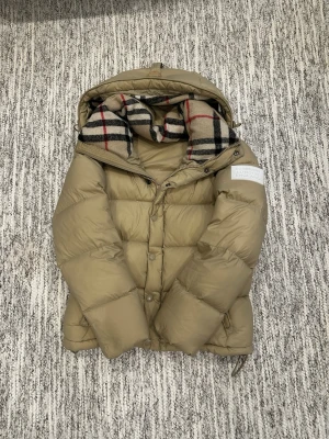 Beige pufferjacka från Burberry - Säljer en beige pufferjacka från Burberry med klassiskt rutigt foder i huvan. Jackan är quiltad, har dragkedja och knappar framtill samt långärmad design. Tillverkad i polyester och syntetmaterial, perfekt för kalla dagar. Its used and condition is 8/10. Its goes perfectly 165-172cm persons. 