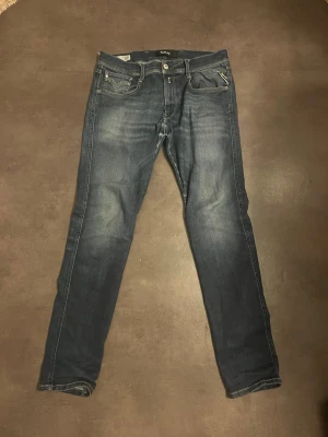 Mörkblå jeans från Replay - Snygga mörkblå jeans från Replay med klassisk femficksdesign och diskreta slitningar framtill. Jeansen har raka ben och tydliga kontrastsömmar. Perfekta för dig som gillar en stilren och tidlös look. 2 små defekter som syns på bilder.