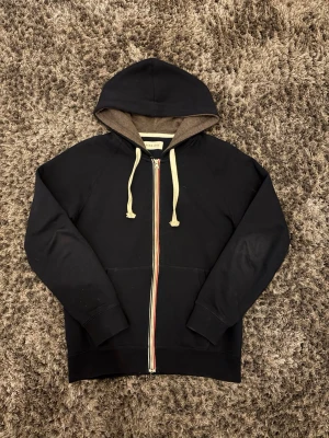 Svart hoodie från Pier One med dragkedja - Svart hoodie från Pier One med hel dragkedja framtill och vita snören i huvan. Insidan av huvan är brunmelerad och dragkedjan har en snygg randig detalj. Perfekt för en chill och avslappnad stil.