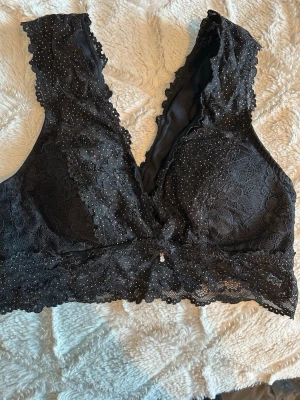 Svart spets-bh med glitterdetaljer - Supersnygg svart bh från Gina Tricot i spets med glittrande detaljer och dekorativ liten rosett framtill. Bh:n har breda axelband och är tillverkad i 90% polyamid och 10% elastan för en bekväm stretchig passform. Perfekt för dig som gillar lite extra bling!