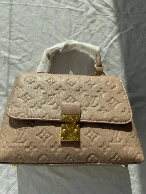 Beige Louis Vuitton handväska - Säljer en beige handväska från Louis Vuitton i präglat läder med det klassiska LV-monogrammet. Väskan har guldfärgade metalldetaljer och ett topphandtag med skyddande tyg. Stängs med ett elegant guldfärgat lås framtill. Perfekt för dig som gillar lyxiga accessoarer.