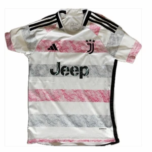 Juventus matchtröja Adidas 23/24 - Juventus matchtröja från Adidas i vitt med breda rosa och gråa ränder. Tröjan har svarta detaljer, tre svarta ränder på axlarna och Juventus-logga med tre stjärnor på bröstet. Jeep-logga på magen och rund hals. Tillverkad i lätt och ventilerande material.
