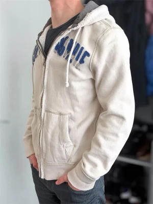 Beige hoodie från Abercrombie - Snygg beige hoodie från Abercrombie med dragkedja, blå broderad text på bröstet och två stora fickor framtill. Mjuk insida och grått foder i huvan. Perfekt för en avslappnad och trendig look.