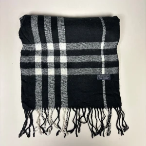 Svartvit rutig halsduk från Burberry - Snygg halsduk i svart och vitt från Burberry med klassiskt rutmönster och fransar i kanterna. Tillverkad i 100% lammull för en mjuk och varm känsla. Perfekt accessoar för att ge din outfit en tidlös touch.
