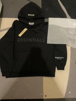 Svart Essentials Fear of God hoodie - Snygg svart hoodie från Fear of God Essentials Oversized passform och mjukt material som känns riktigt skönt. Perfekt för dig som gillar streetwear och vill ha en clean look. Priset kan diskuteras vid snabb affär.