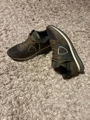 Philippe Model trainers  - Snygga sneakers i mörkgrönt och brunt från Philippe Model. Skorna har mockadetaljer, meshpartier och en vit sula med svart kant. Klassisk låg modell med bruna skosnören och logotyp på sidan. Perfekta för en trendig streetstyle-look.