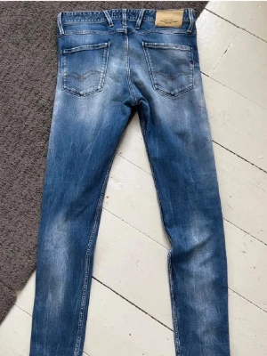 Blå Replay jeans med slitningar - Säljer ett par blå jeans från Replay med snygga slitningar och klassiska bakfickor. Jeansen har en slim fit och är tillverkade i denim med detaljer som läderpatch på linningen. Perfekt för dig som gillar en cool och avslappnad stil.