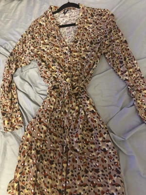 Mönstrad långärmad klänning med knyt - Säljer en långärmad klänning med beige botten och leopardmönster i svart, brun, vit och orange. Klänningen har v-ringning, knappar framtill och ett knytband i midjan för snygg passform. Perfekt för dig som gillar trendiga mönster och bekväma material.
