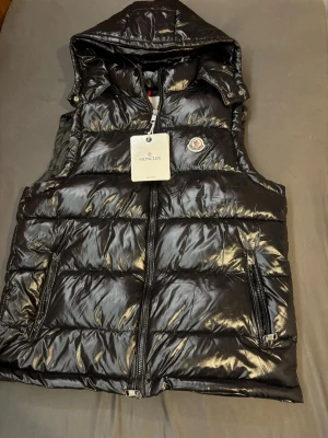 Svart dunväst från Moncler med huva - Snygg svart dunväst från Moncler med glansig finish och avtagbar huva. Västen har två dragkedjeförsedda fickor och klassisk Moncler-logga på bröstet. Perfekt för lager-på-lager och streetwear-stil. Stängs med dragkedja framtill.